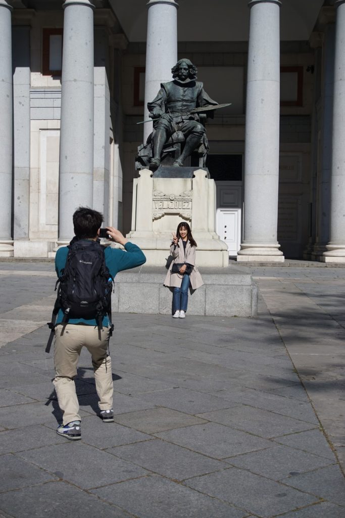A male tourist takes a photo of his female companion seated below a statue of Velazques in front of the Prado museum in Madrid, Spain.